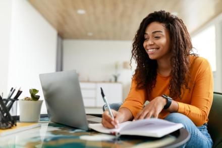 A woman takes notes and smiles while she looks at a laptop screen